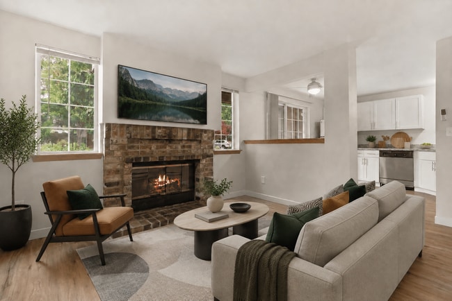 Staged apartment living room with a brick fireplace, cream seating, scenic TV, and an adjacent kitchen with stainless-steel appliances. - Stonegate Apartments