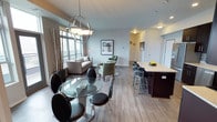 This single-floor residence showcases a thoughtfully designed open-concept layout that seamlessly connects the kitchen, dining, and living areas into one cohesive, airy space. Rich wood-look flooring runs throughout, grounding the interior with a warm yet modern aesthetic.

The kitchen is a standout feature, equipped with dark espresso cabinetry, a horizontal tile backsplash, stainless steel appliances including a French door refrigerator, and a light-toned countertop island with bar seating for two. A double sink and ample upper and lower cabinet storage add to its functionality.

Adjacent to the kitchen, the dining area accommodates a round glass-top table with sleek black chairs, illuminated by a contemporary globe pendant light fixture. Large floor-to-ceiling windows nearby flood the space with natural light and offer expansive outdoor views.

The living area provides a relaxed atmosphere with comfortable seating, including soft sage-green accent chairs and a sofa, complemented by decorative accents. The overall palette of cool grays, deep browns, and soft greens creates a balanced, modern ambiance throughout the residence.