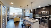 This two-story loft offers a well-designed open-concept layout that seamlessly blends living, dining, and kitchen areas into a cohesive, functional space. The kitchen serves as a centerpiece, featuring dark espresso cabinetry, granite countertops, a stainless steel refrigerator, and a subway tile backsplash. A large kitchen island with a built-in sink doubles as a breakfast bar, complemented by industrial-style adjustable bar stools and elegant pendant lighting that adds a warm, modern ambiance. Rich dark hardwood flooring flows throughout the main level, grounding the space with a sophisticated contrast to the lighter walls and countertops. The living area benefits from expansive floor-to-ceiling windows that flood the interior with natural light, creating an airy and welcoming atmosphere. Recessed lighting and stylish pendant fixtures provide layered illumination throughout. The loft's two-floor configuration adds a sense of dimension and versatility, making excellent use of vertical space. Overall, the space strikes a balance between urban industrial character and contemporary comfort, making it an ideal single-bedroom residence for those seeking modern city living.