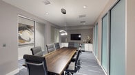 This contemporary conference room is thoughtfully designed to support productive professional gatherings. The centerpiece is a striking live-edge wood conference table, which seats multiple attendees and introduces a natural, organic element that contrasts beautifully with the room's otherwise modern aesthetic. Surrounding the table are high-back ergonomic chairs upholstered in charcoal gray, providing both comfort and a cohesive visual palette.

Overhead, a sculptural pendant light fixture with a layered, ring-style design serves as a focal point, complemented by recessed lighting throughout the ceiling. Neutral gray walls and patterned gray carpet tiles contribute to the room's refined, professional atmosphere.

A wall-mounted flat-screen television anchors the far wall, supported by a sleek white credenza with ample storage. Decorative accents, including ceramic vases and florals, add subtle warmth. Floor-to-ceiling glass panels along one side of the room allow natural light to filter in while maintaining a sense of openness. An adjacent wall features frosted glass partitions, ensuring privacy without sacrificing the airy, contemporary feel of the space.