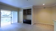 This single-floor space presents a well-appointed living area defined by its clean lines and thoughtful built-in features. The focal point of the main room is a dark-finished built-in unit, comprising open upper shelving and lower closed cabinetry with a sleek countertop surface — ideal for display, storage, or use as a media console. Crown molding runs along the ceiling perimeter, lending a refined architectural detail to the space. Soft, neutral-toned carpeting extends throughout, contributing to a warm and comfortable ambiance underfoot. The walls are finished in a muted warm beige, complementing the dark cabinetry and creating a balanced, cohesive aesthetic. Recessed lighting provides overhead illumination, while vertical blinds along a large sliding door or window allow for adjustable natural light. The doorway visible in the background suggests a connected layout with easy flow between rooms. The space feels open and airy, with ample room for flexible furniture arrangements. Overall, the design balances functional storage solutions with classic interior detailing, resulting in a practical yet polished living environment.