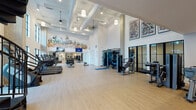 Located at 2395 Herodian Way SE in Smyrna, Georgia, this fitness center is a thoughtfully designed, multi-level facility that combines modern architecture with a comprehensive suite of workout amenities. The main floor is an open, airy space defined by soaring double-height ceilings, exposed ductwork painted white, and pendant lighting that contributes to the bright, energetic atmosphere. Light wood-plank flooring runs throughout, lending warmth to the clean, white interior.

The facility is equipped with an extensive selection of cardio machines, including treadmills and stair climbers positioned along floor-to-ceiling windows that offer views of an outdoor pool courtyard, filling the space with natural light. A dedicated strength training zone features cable machines, weight benches, and a dumbbell rack accompanied by floor-to-ceiling mirrors. A striking, colorful cityscape mural anchors one wall, while abstract artwork adorns the upper walls, adding a creative and dynamic aesthetic.

A dramatic black spiral staircase connects the main floor to an upper mezzanine level, which houses additional equipment, including cycling stations. Black-framed glass partition doors define separate functional zones while maintaining an open, connected feel. A small refreshment station with cabinetry and counter space is conveniently located near the base of the staircase, rounding out this well-appointed fitness destination.