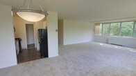 This single-floor space features an open-concept layout that seamlessly connects the living area and kitchen. The spacious living area is finished with neutral-toned wall-to-wall carpeting that adds warmth underfoot, while the clean white walls provide a bright, versatile backdrop throughout. A large window at the far end of the living space allows natural light to filter in, offering pleasant views of surrounding greenery that contribute to a calm, airy ambiance.

The kitchen area is partially visible and transitions from the carpeted living space via a tile floor, offering a practical distinction between zones. A black refrigerator anchors the kitchen, which is partially separated from the living area by a structural wall, providing a sense of defined space while maintaining an open feel. An overhead pendant light fixture with a frosted glass shade is positioned near the kitchen transition, adding a functional and subtle design element. Overall, the space presents a clean, neutral palette that offers a solid foundation for a variety of interior styles and furnishing arrangements.