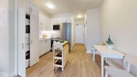 This thoughtfully designed unit offers a smart, single-floor layout comprising a living room, kitchen, and bathroom. Light wood-tone flooring runs continuously throughout, lending warmth and visual cohesion to the entire space.

The living room is bright and inviting, anchored by a large sliding glass door that opens to a private balcony with urban views. The room accommodates a comfortable sectional sofa, area rug, and coffee table, creating a cozy yet functional lounge area.

The kitchen is efficiently arranged in an L-shaped configuration featuring crisp white shaker-style cabinetry, stainless steel appliances including a full-size refrigerator, stove, and microwave. A portable butcher block island provides additional prep space and flexibility. The kitchen flows seamlessly into a dining area, making it ideal for everyday living.

The bathroom is clean and well-appointed, finished in a bright white palette with a vanity featuring under-cabinet storage, a mirror with overhead lighting, and matching light wood flooring. A framed art piece adds a touch of personality to the space.

Throughout the unit, neutral tones and consistent finishes create a cohesive, modern aesthetic that maximizes the sense of openness while maintaining a welcoming, functional atmosphere.