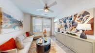 This single-floor residence showcases a thoughtfully designed open layout with consistent light wood-tone flooring and neutral white walls throughout. The living area serves as the heart of the space, featuring a ceiling fan, large windows with horizontal blinds, and a glass door that allows abundant natural light to fill the room. Bold, colorful wall art and a low-profile media credenza add character to the space.

The kitchen is well-equipped with warm-toned wood cabinetry, granite countertops, and a full suite of stainless steel appliances including a gas range, microwave, refrigerator, and dishwasher. A generous peninsula with seating for multiple bar stools provides both additional prep space and casual dining functionality. Recessed lighting illuminates the kitchen efficiently.

The bedrooms are comfortable and well-lit, each featuring ceiling fans, large windows with blinds, and wood-look flooring. One bedroom includes a striking teal upholstered headboard and ample natural light from a corner window arrangement.

The bathroom features warm wood cabinetry, granite countertops, a large vanity mirror with bright overhead lighting, tile flooring, and a shower-tub combination, completing this cohesive and modern residence.