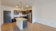 This single-story residence offers a thoughtfully designed open-concept layout where the kitchen flows seamlessly into the living area. The kitchen is well-appointed with dark espresso-toned cabinetry, granite-style countertops, a classic white subway tile backsplash, and a full suite of stainless steel appliances, including a range, microwave, dishwasher, and refrigerator. A central island with matching countertop provides additional workspace and casual dining potential. Elegant brass pendant lights hang above the island, complementing the recessed lighting throughout. The adjoining living area features plush wall-to-wall carpeting and is anchored by a ceiling fan. French doors at the far end of the living space invite natural light and provide access to the outdoors. The bedrooms are carpeted and well-lit, each equipped with a ceiling fan and sizable windows fitted with blinds for light control. The bathroom showcases a sleek, frameless glass-enclosed shower with tiled walls and built-in shelving, paired with a dark-cabineted vanity featuring a granite-style countertop and undermount sink. Light wood-look flooring extends through the kitchen and bathroom areas, creating a cohesive, modern aesthetic throughout the home.