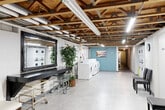 Located at 160 Manitou Ln in Lake Orion, Michigan, this shared laundry facility offers a spacious and functional layout designed to accommodate the needs of multiple residents. The space features an exposed wood truss ceiling with visible ductwork, piping, and fluorescent strip lighting, lending an industrial character while maintaining a clean and organized environment. Large-format light gray floor tiles run throughout, providing a bright, easy-to-maintain surface. The laundry area is well-appointed with multiple front-loading washers and dryers, as well as stacked dryer units, offering ample capacity for residents. A deep utility sink is conveniently positioned near the machines. A long dark-topped folding table runs along one wall, providing generous counter space for sorting and folding laundry. Seating chairs are available for residents waiting on their laundry, and rolling laundry carts are on hand for added convenience. A bulletin board is mounted on the wall for community notices. Decorative touches, including framed wall art and potted plants, add warmth to the utilitarian space. A dedicated closet area with individual storage units is accessible along a corridor at one end of the facility, rounding out the practical amenities available within this well-maintained shared space.