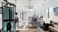 This modern fitness center is designed to support a comprehensive workout experience within a thoughtfully laid-out, single-floor space. The facility is divided into distinct functional zones, each tailored to a specific type of exercise. The cardio area is lined with treadmills and elliptical machines from Matrix, positioned along a windowed wall that allows generous natural light to fill the space and provides views of the surrounding exterior. Ceiling fans circulate air throughout, contributing to a comfortable training environment. A dedicated strength and free weights zone features a dumbbell rack, kettlebell set, and an adjustable weight bench, accompanied by a wall-mounted flat-screen television. A large floor-to-ceiling mirror enhances this area, making the space feel more expansive while allowing users to monitor their form. A cable and multi-functional strength machine rounds out the equipment selection. The flooring transitions between durable hardwood and textured carpet tile, delineating each workout zone. Herringbone-patterned accent tile adds a refined design detail to the white walls. A built-in water station with a bottle-fill fountain and a small storage bench with cubbies offer added convenience. Recessed lighting and ceiling fans complete the clean, contemporary aesthetic throughout.