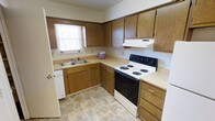 This single-floor space offers a practical and well-organized layout designed for comfortable everyday living. The kitchen serves as a central feature of the home, showcasing warm oak cabinetry that extends from floor to ceiling, providing generous storage throughout. Light-toned laminate countertops run along an L-shaped configuration, offering ample workspace for meal preparation. A double stainless steel sink sits beneath a window that allows natural light to filter in, brightening the room and creating an inviting atmosphere. The kitchen is equipped with a freestanding electric range, complemented by an overhead ventilation hood, making the space both functional and efficient. Durable tile flooring in a neutral earth-tone palette ties the room together while ensuring ease of maintenance. The cabinetry's rich wood finish lends the space a classic, homey character. A dishwasher is also integrated into the layout, adding a modern convenience to the traditionally styled kitchen. Overall, the space presents a clean, no-frills design that prioritizes practicality and utility, making it well-suited for those seeking a straightforward and functional living environment.