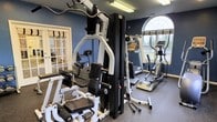 Located at 3998 Audrey Rae Ln in Howell, Michigan, this fitness center offers a well-appointed, single-floor workout facility designed to support a full range of exercise routines. The space is finished with deep navy blue walls and durable rubber flooring, creating a focused and energizing atmosphere suited for physical training.

The facility is equipped with a variety of cardio machines, including multiple elliptical trainers and a treadmill, providing options for low-impact and high-intensity cardiovascular workouts. A multi-station strength training system anchors the space, offering versatile resistance training capabilities for users of all fitness levels. Free weights and additional accessories are also available.

Natural light filters in through several windows, including a striking arched window that adds an architectural accent to the room. The windows also offer views of the surrounding outdoor landscape, contributing to an open and inviting feel despite the dedicated workout environment. Recessed and overhead lighting ensures the space remains well-lit throughout.

A bulletin board and informational postings are mounted on the wall, and a hand sanitizer dispenser adds a practical amenity. The layout is thoughtfully arranged to allow comfortable movement between equipment stations, making this fitness center a functional and well-maintained resource for its users.