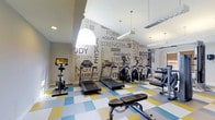 This fitness center is designed to provide a comprehensive workout environment within a single-level layout. The space is immediately eye-catching, thanks to a bold feature wall covered in motivational fitness-related typography, including words such as "Strength," "Endurance," "Flexibility," and "Agility," rendered in varying sizes and shades of gray. The wall serves as both a design statement and an energizing backdrop for workouts.

The flooring consists of a colorful checkered pattern in blue, yellow, and white, adding a dynamic visual element to the space. Overhead, pendant lighting fixtures hang from the ceiling alongside recessed lighting, ensuring the room is well-lit throughout. A large arched window allows natural light to filter in, enhancing the overall brightness of the area.

The equipment lineup includes multiple treadmills, elliptical machines, and a stationary bike, catering to a variety of cardio preferences. A dedicated strength training area is visible along one wall, equipped with a cable machine and weight bench, accommodating resistance and functional training needs. Mirrors along one side of the room amplify the sense of space and allow for form monitoring during exercise.