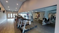 This fitness center offers a spacious, open layout designed to accommodate a comprehensive range of workout activities. The space is divided into distinct functional zones, with a dedicated cardio area prominently featuring a row of treadmills and stationary bikes along one side. The vaulted ceiling in the cardio section adds a sense of openness and airiness, enhanced further by large windows and glass doors that allow generous amounts of natural light to fill the space.

The strength training area is equipped with a variety of cable machines, weight benches, and resistance equipment, thoughtfully arranged to allow comfortable movement between stations. Recessed lighting throughout ensures consistent illumination across all workout zones. The flooring transitions between rubberized gym matting in high-use areas and hard flooring in surrounding sections, providing both functionality and durability.

Motivational artwork and signage are displayed on the walls, contributing to an energetic atmosphere. A support column divides portions of the interior, subtly defining separate workout zones without disrupting the overall open feel. The facility appears well-maintained and thoughtfully organized to serve a diverse range of fitness needs.