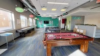 This single-floor clubhouse is designed to serve as a dynamic social and recreational hub. Upon entry, guests are welcomed by a sleek reception area branded with the "Evolve" logo, featuring a clean white desk, a large cylindrical column, and exposed ductwork overhead that lends the space an industrial-modern character. The flooring transitions between wood-look planks and multi-toned carpet tiles with blue and grey accents, adding visual interest throughout.

The expansive game room is a central highlight, equipped with a branded pool table, a foosball table, an air hockey table, a ping pong table, and high-top seating along the windowed walls. Bold teal and dark accent walls energize the area, complemented by decorative pendant lighting and wall-mounted flat-screen televisions.

Multiple lounge zones offer varied seating arrangements, including a curved sectional sofa positioned beside floor-to-ceiling windows that invite abundant natural light and outdoor views. Additional seating areas feature blue modular sofas, leather armchairs, and coffee tables, providing comfortable spaces for relaxing or socializing. The entrance foyer includes a popcorn machine and direct access to the main building, rounding out this well-appointed, amenity-rich clubhouse.