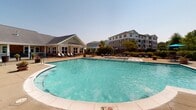 Located at 416 E North Pointe Dr in Salisbury, Maryland, this outdoor amenity area serves as a central gathering space within the community. The centerpiece is a generously sized, freeform resort-style swimming pool with crystal-clear blue water, entry steps with handrails, and in-water lounge chairs for relaxed sunbathing. The pool accommodates both shallow and deeper sections, clearly marked for safety. Surrounding the pool is a spacious concrete sundeck furnished with lounge chairs and patio seating shaded by blue umbrellas, creating a comfortable outdoor retreat. A white pergola structure runs along one side of the deck, providing a shaded walkway that connects to a pool house with restroom and storage facilities. The single-story clubhouse building features a classic design with a pitched shingle roof, stone and siding exterior, covered porch with white columns, and large windows that invite natural light. Decorative planters with seasonal flowers and ornamental iron fencing accent the entryways and transitions between spaces. Mature trees and well-maintained landscaping border the perimeter, offering privacy and a lush green backdrop. The multi-story residential buildings of the surrounding community are visible, providing a sense of the neighborhood's scale and cohesive architectural character.