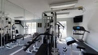 This single-floor fitness center offers a practical and well-appointed workout environment designed to support a full range of exercise routines. The centerpiece of the room is a TuffStuff Apollo 4 multi-station cable machine, providing a comprehensive strength training setup for upper and lower body workouts. Accompanying it are weight benches, a preacher curl bench, and additional resistance training equipment, giving users versatile options for targeted muscle work. A cardio section is visible with at least one elliptical machine, ideal for cardiovascular conditioning. The room features large windows on multiple walls, allowing ample natural light to fill the space and creating an open, energizing atmosphere. Full-length mirrors line one wall, enhancing the sense of space while allowing users to monitor their form during exercise. The flooring is a durable, low-pile carpet suited for a fitness environment, and overhead fluorescent lighting ensures the room remains well-lit throughout. The light gray walls and clean layout contribute to a focused and functional ambiance, making this fitness center a well-rounded facility for daily training needs.