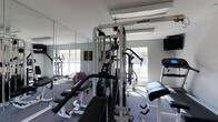 This single-floor fitness center offers a practical and well-appointed workout environment designed to support a full range of exercise routines. The centerpiece of the room is a TuffStuff Apollo 4 multi-station cable machine, providing a comprehensive strength training setup for upper and lower body workouts. Accompanying it are weight benches, a preacher curl bench, and additional resistance training equipment, giving users versatile options for targeted muscle work. A cardio section is visible with at least one elliptical machine, ideal for cardiovascular conditioning. The room features large windows on multiple walls, allowing ample natural light to fill the space and creating an open, energizing atmosphere. Full-length mirrors line one wall, enhancing the sense of space while allowing users to monitor their form during exercise. The flooring is a durable, low-pile carpet suited for a fitness environment, and overhead fluorescent lighting ensures the room remains well-lit throughout. The light gray walls and clean layout contribute to a focused and functional ambiance, making this fitness center a well-rounded facility for daily training needs.