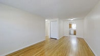 This single-floor space offers a clean, straightforward layout designed for everyday functionality. The main living area is generously sized, featuring light-toned hardwood floors that run throughout, lending warmth and continuity to the space. Crisp white walls and a textured ceiling contribute to the bright, airy atmosphere, while flush-mount ceiling lighting ensures consistent illumination throughout.

A built-in closet with white panel doors provides convenient storage within the main living area, while an open passageway leads to additional rooms beyond, suggesting a practical and connected floor plan.

The kitchen is visible from the main living space and features wooden cabinetry, a window that allows natural light to filter in, and standard appliances including a dishwasher. The light-toned flooring continues seamlessly into this area, maintaining a cohesive look throughout.

Overall, the space presents a neutral, move-in-ready aesthetic with simple finishes that offer a versatile foundation, making it well-suited for a variety of furnishing and décor styles.