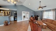 Located at 3414 Shortcut Rd in Pascagoula, Mississippi, this single-story home offers an open, well-connected layout finished with warm terracotta tile flooring that flows consistently throughout the main living areas. The living room is anchored by dramatic vaulted ceilings and ceiling fans, creating a bright and airy atmosphere. Comfortable seating, a wall-mounted television, and a cozy area rug define the space, while a built-in desk area along one wall provides a functional workspace. A set of classic French doors connects the living room to an adjacent sunlit room, enhancing the sense of openness. The dining area is generously sized to accommodate a large eight-seat wooden table and benefits from abundant natural light streaming through multiple windows and two sets of French doors that open directly to an outdoor pool area, offering pleasant views of the surrounding greenery. The cool blue-gray wall color used throughout the home lends a calm, cohesive tone to every room. Recessed lighting and ceiling-mounted fans are found across the interior, ensuring both comfort and practicality. The combination of vaulted ceilings, natural light, and seamless indoor-outdoor connectivity gives this home a relaxed yet functional character well-suited to everyday living and entertaining.