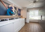 Located on E Roxboro Road NE in Atlanta, Georgia, this single-floor amenity facility provides residents with a well-equipped shared laundry room and a dedicated package locker area. The laundry room features a row of commercial-grade front-load washers and dryers lined along one wall, complemented by a continuous butcher-block folding counter that offers ample workspace. A stacked washer and dryer unit near the entrance provides additional laundry capacity. Warm terracotta tile flooring runs throughout the space, creating a clean and cohesive look, while recessed ceiling lighting ensures bright, even illumination. A playful large-format mural with the "Loads of Fun!" theme adds a lively, welcoming character to the room. Wooden bar stools positioned along the counter provide a convenient spot to wait. The adjacent package locker area is outfitted with a full bank of multi-sized, secure parcel lockers—ideal for receiving deliveries at any hour. A window with blinds allows natural light to filter into this section, adding a sense of openness. Framed wall art near the entryway and well-maintained, neutral-toned walls contribute to the tidy, functional ambiance of the overall space.