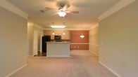 This single-floor space showcases an open-concept layout that seamlessly connects the living area and kitchen, creating a spacious and functional environment. The living area is finished with plush carpeting and neutral-toned walls, complemented by crown molding along the ceiling perimeter. A ceiling fan with an integrated light fixture serves as a practical centerpiece overhead, providing both airflow and ambient lighting.

The kitchen is equipped with light wood cabinetry, a black refrigerator, and a built-in microwave. A peninsula-style island with a light stone countertop offers additional workspace and acts as a natural divider between the kitchen and living areas. Pendant lights hang above, providing task lighting over the island and dining space.

A striking terracotta-toned accent wall adds a warm, distinctive visual element to the open dining area, which is further highlighted by a decorative pendant light. The kitchen features hardwood-style flooring, while the adjoining living space transitions to carpeting. The overall ambiance is warm and inviting, with a cohesive design that balances functionality and comfort throughout the open floor plan.