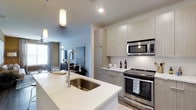 This contemporary single-floor residence showcases a thoughtfully designed open-concept layout that seamlessly connects the kitchen and living areas. The kitchen is a standout feature, outfitted with stainless steel appliances including a range, microwave, and a deep undermount sink set into an expansive quartz waterfall island. Flat-panel cabinetry in a light wood finish extends to the ceiling, maximizing storage while maintaining a clean, minimalist aesthetic. Pendant lighting with warm-toned fixtures hangs above the island, adding an inviting ambiance to the space. The kitchen transitions naturally into the living area, which benefits from abundant natural light streaming through large windows dressed with patterned curtains. The living space offers a relaxed yet refined atmosphere, anchored by a media area and complemented by smooth flooring throughout. Recessed lighting and a neutral color palette of soft whites and warm grays tie the entire space together, creating a cohesive and modern feel. The overall design balances functionality and style, making this residence an ideal space for both everyday living and entertaining.