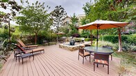 This thoughtfully designed outdoor space offers a serene and functional retreat centered around a spacious wood deck and an intricate network of stone pathways. The deck serves as the primary gathering hub, furnished with rattan dining sets topped by large market umbrellas, as well as open bench seating for relaxed lounging. At the heart of the deck sits a stone-framed fire pit with a decorative water feature, creating a striking focal point that adds both visual interest and ambient sound.

Curved flagstone walkways meander through richly planted garden beds, bordered by manicured boxwood hedges, ornamental grasses, and mature trees that provide generous shade and privacy. Towering bamboo groves add a dramatic, architectural quality to the planting scheme, while layered shrubs and ground cover create a lush, verdant atmosphere throughout.

String lights are strung overhead, enhancing the ambiance during evening hours. A dedicated outdoor kitchen and grill station expands the entertainment potential of the space. The surrounding multi-story building features private balconies that overlook the courtyard below, further integrating indoor and outdoor living. Overall, this outdoor environment is designed to encourage relaxation, social gathering, and a strong connection to nature.