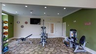 This fitness center offers a functional and inviting workout environment designed to support a range of exercise routines. The space is anchored by professional-grade Hoist strength training equipment, including a multi-purpose cable machine and a dedicated rowing/resistance machine, alongside an adjustable weight bench — providing comprehensive options for both strength and conditioning workouts. A wall-mounted flat-screen television adds an entertainment element, making workouts more engaging. The room features a distinctive patterned carpet with circular motifs in neutral tones, adding visual interest underfoot. Recessed lighting is evenly distributed across the ceiling, ensuring the space is well-lit throughout. Crown molding details lend a polished, finished look to the room. An accent wall painted in a bold olive green creates a dynamic focal point, contrasting with the clean white walls and ceiling. Decorative signage adds personality and character to the space. Access is provided through a paneled white door, and the layout allows ample room to move freely between equipment. Overall, this single-floor fitness center balances practicality with a welcoming atmosphere suited for daily use.