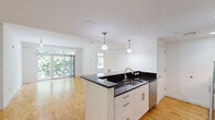 Located at 7 Cameron Ave in Cambridge, MA, this two-bedroom, one-bathroom residence offers a well-designed single-floor layout with a clean, modern aesthetic throughout. The open-concept kitchen and living area serves as the heart of the home, featuring warm hardwood flooring, a central kitchen island topped with dark granite countertops, white shaker cabinetry, and stainless steel appliances including a refrigerator, microwave, and range. Pendant lighting above the island adds a refined touch. The living area extends toward a wall of floor-to-ceiling sliding glass doors that open to a private balcony, flooding the space with natural light and offering lush, tree-lined views. Both bedrooms are carpeted and feature large windows that frame verdant outdoor scenery, creating bright and comfortable retreats. The primary bedroom includes a closet and direct access to the full bathroom. The bathroom is finished with white tile walls, mosaic tile accents, dark granite vanity countertops, white cabinetry, and large-format tile flooring. A connecting hallway ties the layout together efficiently. The overall ambiance is bright, clean, and contemporary, making this a well-appointed residence in a desirable Cambridge location.