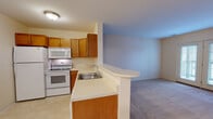 Located at 1900 Sweet Home Rd in Buffalo, New York, this single-floor unit offers a functional and well-organized layout. The kitchen is equipped with warm honey-toned wood cabinetry, light countertops, and white appliances including a refrigerator, electric range, and over-the-range microwave. A peninsula with a built-in sink and breakfast bar separates the kitchen from the living area, creating an open-concept flow. The accent wall in a soft blue hue adds a subtle visual distinction between the two spaces. The living area features neutral carpeting and is bathed in natural light, with French doors leading to a private balcony overlooking the exterior. The bedroom is a generously sized, carpeted room with a window fitted with blinds, providing natural light and outdoor views. The bathroom is clean and practical, featuring tile flooring, a wood-toned vanity with a single sink, a well-lit mirror, and a glass-enclosed shower stall. Throughout the unit, the consistent use of neutral tones, light countertops, and wood accents creates a cohesive and inviting atmosphere suited for comfortable everyday living.