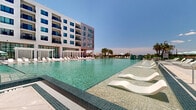 Located at 4311 34th St S in Saint Petersburg, Florida, this expansive amenity level showcases a thoughtfully designed outdoor environment that blends resort-style recreation with modern residential architecture. The space is anchored by a large, sparkling swimming pool with tiled steps, surrounded by rows of white lounge chairs and shaded cabanas, creating an inviting atmosphere for relaxation under the Florida sun.

Adjacent to the pool, a lush green lawn provides open gathering space with umbrella-shaded dining tables and seating, ideal for casual outdoor socializing. The lawn areas are framed by well-maintained planters and tropical greenery, adding softness to the clean, contemporary aesthetic of the surrounding multi-story white building with its grid-pattern balconies and large dark-framed windows.

A covered porte-cochère entrance features warm wood-panel ceilings, ceiling fans, and cylindrical columns alongside wicker lounge seating, offering a welcoming and shaded arrival experience. An elevated sun terrace provides additional lawn seating with lounge chairs, open-air views of the surrounding landscape, and railing-framed panoramas stretching toward the horizon.

Throughout the amenity level, polished concrete pathways connect each zone seamlessly, while the interplay of sunlight, open sky, and thoughtfully placed shade structures creates a balanced, resort-caliber outdoor living environment.