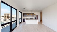 This contemporary single-floor residence showcases a clean, modern design throughout, defined by light hardwood flooring, crisp white walls, and bold black-framed floor-to-ceiling windows that flood every room with natural light and frame urban skyline views. The open-concept kitchen is a standout feature, equipped with a two-tone cabinet scheme pairing white uppers with dark wood lowers, a striking marble-look backsplash, a central island with a dark countertop, stainless steel appliances, and gold pendant lighting that adds warmth to the space. A gold-tone faucet and integrated sink complete the refined aesthetic. Multiple bedrooms offer generous proportions, each with large sliding glass doors or oversized windows that provide city and treetop views, along with light hardwood floors and clean white finishes. Walk-in closet space with wire shelving systems provides practical storage. The bathrooms are finished with dark large-format tile flooring and white cabinetry. The primary bath features a floating double-sink vanity with gold fixtures and a backlit mirror, while a secondary bath offers a soaking tub and shower combination with gold hardware and subway tile surround. Throughout the residence, a consistent palette of white, gold, and natural wood creates a cohesive and polished contemporary living environment.