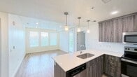 Located at 308 W North Temple St in Salt Lake City, Utah, this 690-square-foot apartment offers a practical and modern single-floor layout with 2 bedrooms and 1 bathroom. The open-concept kitchen and living area serves as the centerpiece of the unit, featuring a large island with a light stone countertop, undermount sink, and pendant lighting overhead. Dark wood-grain cabinetry extends floor-to-ceiling, complemented by stainless steel appliances including a refrigerator, range, and microwave. Light wood-style flooring runs throughout the main living area, which receives natural light through a bank of windows fitted with horizontal blinds and includes a door with a sidelight panel providing outdoor access. The primary bedroom measures 140 square feet and includes double closets and a ceiling fan. The second bedroom also features carpeted flooring, a window with outdoor views, and a double closet. Both bedrooms offer neutral walls and carpet. The shared bathroom includes a tub-shower combination, vanity, and wood-style flooring. A hallway connects the rooms and provides access to in-unit laundry. The unit's clean, neutral palette and modern finishes contribute to a cohesive overall aesthetic.