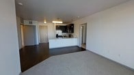 This single-floor space showcases a contemporary open-concept design that integrates the kitchen and living areas into one cohesive environment. The kitchen is equipped with stainless steel appliances, including a refrigerator and range, complemented by dark upper cabinetry that contrasts sharply against crisp white lower cabinetry and a light granite-style countertop. A peninsula with seating potential extends from the kitchen, offering a natural divide between the cooking and living spaces. Pendant lighting above the peninsula and a flush ceiling fixture provide focused illumination throughout. The flooring transitions from dark hardwood-style material in the kitchen zone to neutral, plush carpeting in the living area, adding textural variety. The walls are finished in a clean, neutral white tone, enhancing the sense of openness. Multiple doorways are visible, suggesting access to additional rooms such as a bathroom and a separate enclosed space. The overall ambiance is modern and functional, with a clean aesthetic defined by strong color contrasts and practical design choices.