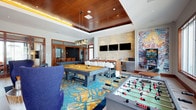 This single-story clubhouse and entertainment lounge is designed for social interaction and recreation, combining multiple distinct areas within a cohesive, upscale aesthetic. The main game room serves as a central hub, featuring a pool table, foosball table, and shuffleboard set atop a bold teal-and-gold botanical-patterned carpet. A striking coffered ceiling clad in warm wood panels with integrated ambient lighting anchors the space visually. A wet bar area with a stone-tile accent wall, bar-height seating, beverage refrigerators, and multiple mounted flat-screen televisions provides a focal point for entertainment. Vibrant large-scale artwork and a decorative pinball machine add personality and color throughout. Adjacent to the game room, a comfortable lounge area centers on a linear gas fireplace set within a floor-to-ceiling stacked-stone wall, flanked by plush navy wingback chairs and modern accent furniture. Large wood-framed windows and glass doors invite natural light throughout. A dedicated private screening room features rows of tiered, cognac-leather reclining theater seats atop coordinating patterned carpet, with dark walls and subtle sconce lighting creating an immersive cinematic atmosphere. Together, the areas offer a well-appointed, resort-caliber amenity space.