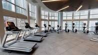 This high-end fitness center is defined by its striking architectural design, featuring soaring floor-to-ceiling windows that wrap around much of the space, flooding the interior with natural light and framing sweeping urban skyline views. The facility is organized across several distinct zones, each designed to support a comprehensive workout experience. The main cardio area is equipped with a row of modern treadmills, elliptical trainers, and stationary bikes, all positioned to take full advantage of the panoramic city views. The curved ceiling in this section is accented with sleek linear lighting and warm wood paneling, lending a refined, contemporary aesthetic. A dedicated functional training zone houses a punching bag, TRX suspension systems, medicine balls, stability balls, resistance bands, and step platforms, providing versatile options for dynamic workouts. The free weights area includes a well-stocked dumbbell rack and additional strength equipment, positioned against another wall of large windows. A separate studio space with light hardwood flooring, sheer curtains, and soaring ceilings offers an airy, open environment ideal for cycling classes or personal training. Durable rubber flooring throughout the main workout zones ensures both safety and performance. The facility connects seamlessly between areas via glass-door transitions, maintaining an open and cohesive feel throughout.