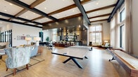 Located at 1400 N Main St in Keller, Texas, this single-floor clubhouse is a generously scaled community gathering space designed with a sophisticated, contemporary aesthetic. The open-plan layout is anchored by striking exposed dark wood beams set against crisp white ceilings, creating a warm yet modern atmosphere throughout.

The main gathering area features light hardwood-style flooring and is furnished with distinct zones for socializing and recreation, including a full-size ping pong table as a central focal point. A built-in lounge nook showcases a sleek fireplace flanked by a built-in shelving unit with display space and an overhead flat-screen TV, providing a cozy retreat within the larger space.

A prominent bar area serves as a social hub, complete with a granite-topped counter, upholstered bar seating, dramatic gold pendant lighting, and a decorative arched shelving display backed by dark cabinetry. Adjacent to this is a dining area with ample seating for group gatherings.

Floor-to-ceiling windows dressed in sheer linen curtains flood the space with natural light and offer views of the surrounding community. A dedicated reception area features marble-accented counters and dark wood paneling, while glass-walled private rooms provide flexible meeting or office space. The overall ambiance is refined, welcoming, and well-appointed throughout.