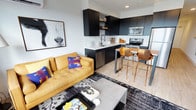 This stylish, single-floor space showcases a thoughtfully designed open-concept layout where the living area and kitchen flow seamlessly together. The kitchen is outfitted with floor-to-ceiling dark matte cabinetry that creates a bold, contemporary contrast against the light walls and white countertops. Stainless steel appliances, including a range, microwave, dishwasher, and refrigerator, provide a polished, functional finish, while open shelving with warm wood accents adds an inviting touch. A sleek breakfast bar with copper-toned sculptural bar stools serves as a natural divider between the kitchen and living area, offering a casual dining option.

The living area is anchored by a rich mustard-yellow leather sofa adorned with vibrant accent pillows, set atop a graphic black-and-white area rug. A concrete-style coffee table grounds the space, while a large, striking black-and-white photographic artwork on the wall serves as a bold focal point. Light hardwood flooring runs throughout, enhancing the sense of openness and warmth. A wall-mounted mini-split unit and recessed lighting ensure year-round comfort. The overall ambiance is modern and eclectic, blending bold colors with industrial-inspired design elements.