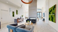 This single-floor amenity space in California's 95330 zip code presents a thoughtfully designed community hub with a cohesive modern aesthetic. The centerpiece is a striking main lobby and lounge featuring soaring double-height ceilings anchored by a dramatic floor-to-ceiling fireplace surround in muted gray. A sculptural copper ring chandelier draws the eye upward, while terrazzo flooring — consistent throughout the entire space — adds texture and visual continuity. Velvet lounge seating is arranged around a wooden coffee table, and a reception desk with warm wood accents sits adjacent to living moss wall art. Glass doors at the rear of the lobby provide direct views to an outdoor pool area, flooding the space with natural light.

A dedicated conference room features a large chevron-patterned dark wood table surrounded by leather chairs, pendant lighting, and windows with neighborhood views. Adjacent is a well-appointed kitchenette with white cabinetry, a stone countertop, and a full-height iridescent teal tile accent wall that serves as a bold focal point.

A casual lounge or game room offers a relaxed retreat with a sectional sofa, natural light, and direct outdoor access. A clean corridor with terrazzo floors connects the various zones, completing this polished and functional communal environment.
