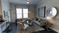This single-floor space showcases a thoughtfully designed living area that blends modern aesthetics with comfortable functionality. The living room is anchored by polished dark concrete-style flooring, which contrasts beautifully against the neutral gray walls and light-toned ceiling, creating a sophisticated urban atmosphere. A ceiling fan with a built-in light fixture serves as a practical centerpiece overhead, complemented by crown molding that adds a refined architectural detail.

Large windows dressed with white plantation shutters and floor-length draping panels allow generous natural light to filter into the space, lending an open and airy feel. The room is furnished with a contemporary gray sofa adorned with decorative pillows in muted and olive tones, paired with a sleek chrome-framed white leather lounge chair. A geometric mirrored coffee table anchors the seating arrangement atop a plush, textured area rug. A dramatic arc floor lamp provides warm ambient lighting, while a large abstract canvas artwork adds a bold visual statement to one wall. The overall ambiance is polished, modern, and inviting.