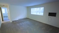 This single-floor space presents a clean, straightforward layout centered around a generously sized main living area. The room is finished with wall-to-wall carpeting in a neutral tone, providing a practical and comfortable underfoot surface that complements the light-colored walls throughout. A large window positioned along one wall serves as a key architectural feature, allowing natural light to fill the interior while offering a view of the surrounding greenery outside. Horizontal blinds on the window provide adjustable light control and privacy. Heating is provided by a baseboard unit installed along the lower portion of the wall beneath the window, a functional and unobtrusive climate-control solution. The walls are painted in a soft, neutral off-white tone, contributing to a bright and open atmosphere. The ceiling is standard height, maintaining a simple and clean aesthetic that suits the overall utilitarian character of the space. The room's open, unfurnished layout allows for flexible furniture arrangements to suit a variety of uses. Overall, the space offers a modest yet functional environment with practical finishes and adequate natural light.