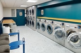 The Pelham Hall Laundry facility is a well-organized, single-floor space spanning 260 square feet. The room is designed for maximum efficiency, featuring two rows of commercial-grade washers and dryers along the walls. The machines are stainless steel, giving the space a modern and clean appearance.

The color scheme is striking, with teal blue walls contrasting against white ceilings and terrazzo-style flooring. Yellow accents, visible on countertops and trim, add a cheerful touch to the space. Bright, recessed lighting fixtures in the drop ceiling ensure excellent visibility throughout the area.

Practicality is key in this laundry room. Long countertops above and below the machines provide ample space for folding and organizing laundry. Built-in benches with cushions offer seating for those waiting on their laundry cycles. The room also includes trash receptacles and possibly detergent vending machines, though these are not clearly visible in the images.

Artistic elements soften the utilitarian nature of the space. Abstract artwork adorns the walls, adding visual interest and a touch of sophistication. An exit sign and fire alarm system are prominently displayed, ensuring safety compliance.

The layout is designed for easy navigation, with a clear pathway between machine rows. An adjoining hallway suggests this laundry facility is part of a larger building complex, likely serving multiple residents or students. Overall, the space balances functionality with aesthetic appeal, creating a pleasant environment for a typically mundane task.