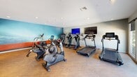 This state-of-the-art fitness center, situated at 11152 Wallingsford Rd in Los Alamitos, CA, spans 970 square feet on a single floor. The facility is thoughtfully divided into distinct workout zones, each equipped with professional-grade exercise machines. The cardio area features a variety of equipment including treadmills, ellipticals, and stationary bikes, all facing large windows that flood the space with natural light and offer views of an outdoor pool area. A separate strength training section is outfitted with weight machines and free weights, complemented by a spacious area for floor exercises and stretching. The center boasts high ceilings with recessed lighting, creating an open and airy atmosphere. Wood-look flooring throughout adds warmth to the space while providing durability for heavy equipment. Walls painted in a combination of neutral tones and vibrant accents create a motivating environment. Large mirrors line some walls, enhancing the sense of space and allowing users to monitor their form. Amenities such as water stations and towel service points are strategically placed for convenience. The layout promotes easy circulation and accessibility, making it an inviting space for fitness enthusiasts of all levels.