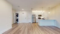 This well-appointed single-floor residence offers a clean, modern aesthetic defined by light wood-look flooring, recessed lighting, and a neutral warm-toned palette that flows seamlessly throughout every room.

The open-concept living and kitchen area serves as the heart of the unit. The kitchen is outfitted with white shaker-style cabinetry, stainless steel appliances including a refrigerator, electric range, and over-the-range microwave, as well as a white subway tile backsplash and a peninsula island with pendant lighting above — ideal for both cooking and casual dining. A dedicated in-unit closet houses a stacked washer and dryer, adding a practical and convenient touch near the main entry.

The living area is generously proportioned and filled with natural light from large windows and a glass door that opens to an outdoor space, offering views of the surrounding community. A wall-mounted mini-split unit ensures climate control comfort year-round.

The bedroom is a calm, light-filled retreat with a large window framing exterior views, while the bathroom impresses with a floor-to-ceiling marble-style tiled walk-in shower enclosed by a frameless sliding glass door, a white vanity with clean countertops, and coordinating wood-look tile flooring. The overall design of this residence strikes a balance between contemporary style and functional living.