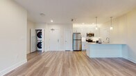 This well-appointed single-floor residence offers a clean, modern aesthetic defined by light wood-look flooring, recessed lighting, and a neutral warm-toned palette that flows seamlessly throughout every room.

The open-concept living and kitchen area serves as the heart of the unit. The kitchen is outfitted with white shaker-style cabinetry, stainless steel appliances including a refrigerator, electric range, and over-the-range microwave, as well as a white subway tile backsplash and a peninsula island with pendant lighting above — ideal for both cooking and casual dining. A dedicated in-unit closet houses a stacked washer and dryer, adding a practical and convenient touch near the main entry.

The living area is generously proportioned and filled with natural light from large windows and a glass door that opens to an outdoor space, offering views of the surrounding community. A wall-mounted mini-split unit ensures climate control comfort year-round.

The bedroom is a calm, light-filled retreat with a large window framing exterior views, while the bathroom impresses with a floor-to-ceiling marble-style tiled walk-in shower enclosed by a frameless sliding glass door, a white vanity with clean countertops, and coordinating wood-look tile flooring. The overall design of this residence strikes a balance between contemporary style and functional living.