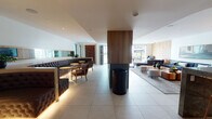 Located at 8809 Washington Blvd in Culver City, California, this single-floor space is a thoughtfully designed communal lounge that blends modern aesthetics with comfortable functionality. The open-plan layout is anchored by large-format porcelain tile flooring that flows seamlessly throughout, connecting distinct seating zones that cater to both social gatherings and relaxed lounging.

One area features richly upholstered tufted booth seating in deep charcoal, paired with warm-toned accent chairs and gold bistro tables, accented by an exposed brick backdrop and ambient under-shelf lighting. Adjacent to this, a more relaxed lounge zone showcases a generous L-shaped navy sectional sofa, complemented by a mixed-wood coffee table and a framed urban artwork displayed against light wood panel wall cladding. Natural light pours in through expansive windows, offering views to the outdoors and adding brightness to the crisp white ceilings.

The kitchenette is a design highlight, featuring light oak cabinetry with brass hardware, a sculptural patterned tile backsplash in muted blue-grey tones, stone countertops, and a row of teal pendant lights that add a distinctive pop of color. Equipped with a stainless steel sink, under-counter refrigeration, and warming drawers, the kitchenette is as practical as it is visually striking. Together, these elements create a refined, welcoming environment ideal for community use or professional gatherings.