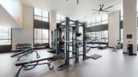 This expansive, single-floor fitness center is a well-appointed facility designed to support a full range of workout routines. The space is defined by its striking double-height ceilings and floor-to-ceiling windows that flood the interior with natural light while offering panoramic urban skyline views — creating an energizing atmosphere throughout.

The main workout floor features durable speckled rubber flooring and is equipped with multiple power racks and squat stations outfitted with Olympic barbells and weight plates, ideal for strength training. A dedicated section houses a full lineup of cardio machines, including treadmills, ellipticals, and stair climbers, all positioned to take advantage of the city views. Cable machines, adjustable benches, and a comprehensive free weights area with multiple dumbbell racks round out the strength training offerings.

A separate studio room with warm hardwood flooring accommodates cycling bikes and stretching or group fitness activities, with full-length mirrors and additional dumbbells. Large ceiling fans circulate air throughout the space. A motivational wall mural with marble-style accents and the phrase "Forward Is The Way" adds a bold, design-forward touch. The facility is thoughtfully zoned to serve both high-intensity training and focused individual workouts.