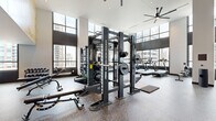 This expansive, single-floor fitness center is a well-appointed facility designed to support a full range of workout routines. The space is defined by its striking double-height ceilings and floor-to-ceiling windows that flood the interior with natural light while offering panoramic urban skyline views — creating an energizing atmosphere throughout.

The main workout floor features durable speckled rubber flooring and is equipped with multiple power racks and squat stations outfitted with Olympic barbells and weight plates, ideal for strength training. A dedicated section houses a full lineup of cardio machines, including treadmills, ellipticals, and stair climbers, all positioned to take advantage of the city views. Cable machines, adjustable benches, and a comprehensive free weights area with multiple dumbbell racks round out the strength training offerings.

A separate studio room with warm hardwood flooring accommodates cycling bikes and stretching or group fitness activities, with full-length mirrors and additional dumbbells. Large ceiling fans circulate air throughout the space. A motivational wall mural with marble-style accents and the phrase "Forward Is The Way" adds a bold, design-forward touch. The facility is thoughtfully zoned to serve both high-intensity training and focused individual workouts.