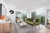 This thoughtfully designed single-floor residence combines modern finishes with warm, eclectic décor to create a welcoming and functional living environment. The entryway sets the tone with light wood flooring, crisp white walls, and curated artwork, leading seamlessly into the main living spaces.

The kitchen is a standout feature, outfitted with sage-green and blue cabinetry, stainless steel appliances, and a clean subway tile backsplash. A large sliding glass door floods the space with natural light and opens to an outdoor balcony with treetop views, complemented by a natural wood dining table nearby.

The living room is bright and open, anchored by floor-to-ceiling sliding glass doors that provide direct balcony access and sweeping outdoor views. The space comfortably accommodates seating and lounging areas, creating a relaxed atmosphere.

Two well-proportioned bedrooms each feature light wood flooring, natural light from large windows or sliding glass doors, and access to the outdoors. Both bathrooms mirror each other in design, featuring grey cabinetry with white countertops, warm wood flooring, and backlit vanity mirrors for a clean, contemporary aesthetic.

Throughout the residence, light wood flooring, white walls, and large windows create a cohesive, airy ambiance that balances modern design with comfortable living.