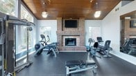 This fitness center combines functional design with inviting architectural character. The space is distinguished by a warm, rich wood-paneled ceiling accented by pendant lighting, which adds a refined aesthetic rarely found in a gym environment. A full brick fireplace serves as a striking focal point, complete with a wall-mounted flat-screen television above the mantle, creating an engaging atmosphere for workouts.

The facility is generously lined with large windows and floor-to-ceiling glass panels that flood the interior with natural light and offer views of a lush outdoor area, including what appears to be a pool and patio space. High vaulted ceilings in certain sections further enhance the sense of openness. The walls feature a two-tone design with blue and white accents, complemented by full-length mirrors that visually expand the space and assist with form during exercise.

Rubber flooring throughout provides both safety and durability. The equipment selection is comprehensive, including multiple Cybex treadmills, elliptical trainers, stationary bikes, a rowing machine, and a cable strength training machine. A beverage vending machine is also conveniently located near the entry. The overall ambiance strikes a balance between a motivating workout environment and a comfortable, well-designed interior.
