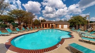 This outdoor pool facility in Jackson, Mississippi, is designed to offer a comfortable and functional recreational experience. The centerpiece is a generously sized freeform swimming pool with vivid turquoise water, framed by brick coping that adds a classic, durable aesthetic. Depth markers are clearly visible, and the pool's curved shape provides an inviting, resort-style appearance.

Surrounding the pool is an expansive concrete sundeck furnished with teal lounge chairs arranged along the perimeter, offering ample space for relaxation. Shaded seating areas with striped patio umbrellas and coordinating teal chairs and tables provide comfortable spots for those seeking relief from the sun. Mature trees line the fenced boundary, offering natural shade and privacy to the space.

A single-story brick clubhouse anchors one side of the pool area, featuring a pitched roof, white-trimmed windows, and a garage-style door, likely housing maintenance or storage facilities. Adjacent to the clubhouse, a covered porch with sturdy brick columns and a beadboard ceiling extends along the building, accessed through white French doors. The porch is enclosed by wrought iron fencing and gating, reinforcing the secure, well-maintained character of the overall facility.